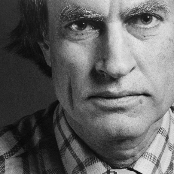 Closeup of a man with a serious expression looking into the camera and wearing a checked shirt.