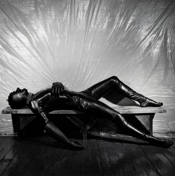 Man dressed in a head-to-toe rubber suit lying on his back on wooden bench in front of a plastic tarp which forms a halo around his body.