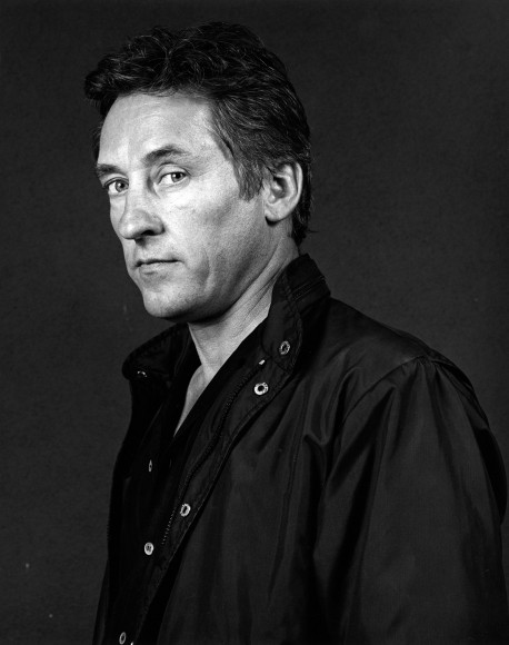 The artist Ed Ruscha in three-quarter view with his eyes turned to look at the viewer and wearing a black windbreaker.