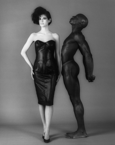 A woman in a leather cocktail dress and high heels facing the camera. An almost nude man stands next to her facing her and looking upward.