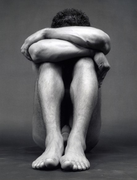 Black male nude sitting down, hands covering face and feet together.