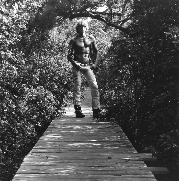 Shirtless white male with hands on hips, standing on boardwalk surrounded by foliage.