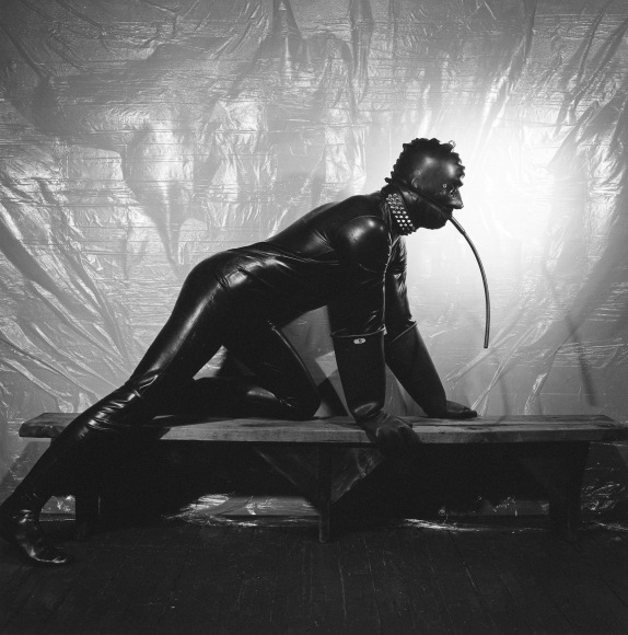 Man in rubber suit on all fours on wooden bench.