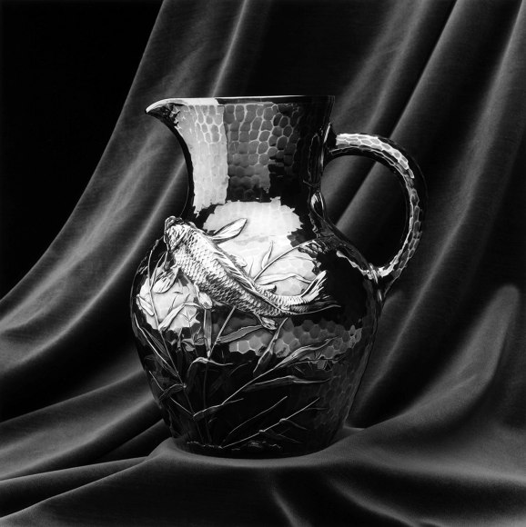 Silver pitcher with a fish design against a draped cloth background.