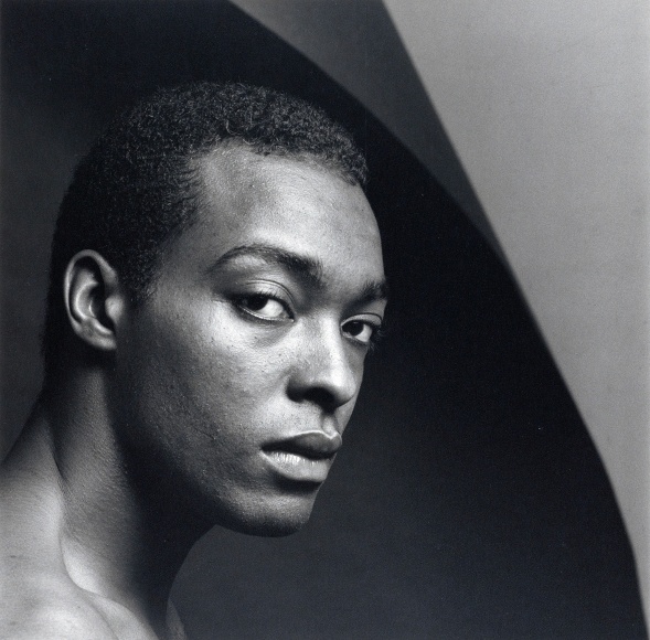 Close-up of shirtless male turning his gaze toward the viewer in front of a curved geometric background.