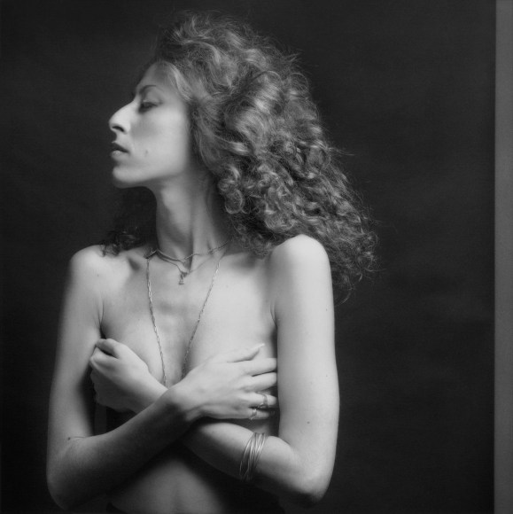 Portrait of a topless woman with curly hair and silver jewelry turning her face in profile and crossing her arms to cover her breasts.