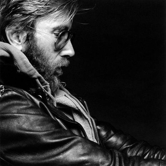 Close-up profile view of a man with a beard wearing sunglasses and a leather jacket over a zip-up sweatshirt.