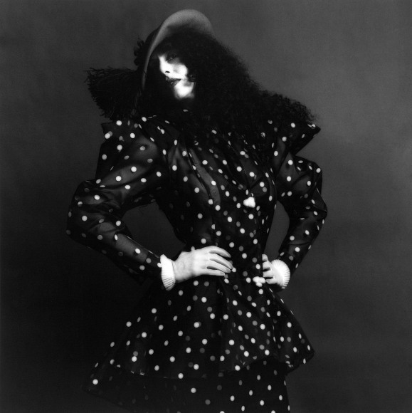 Woman wearing a polka dot dress and hat stands with her arms on her hips looking at the viewer.