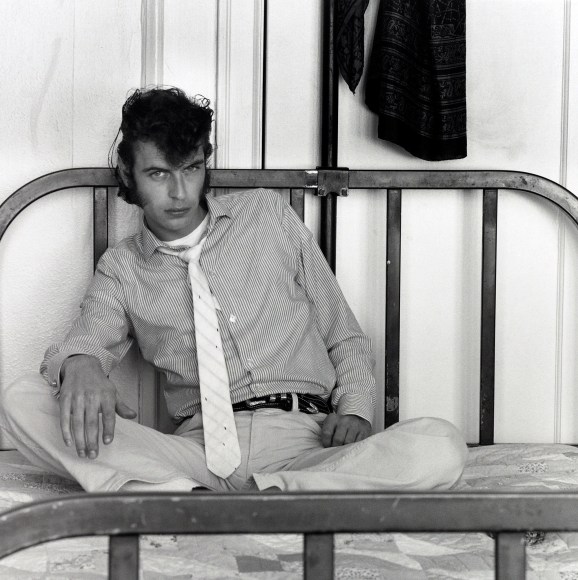 Man sitting cross-legged against the headboard of a bed looking at the camera.