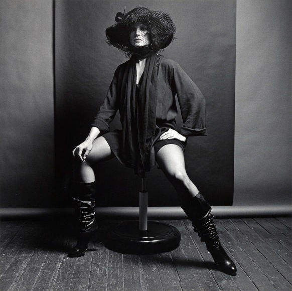 Woman wearing a veiled hat, tunic, and knee-high leather boots sitting on a stool in front of a black and grey backdrop.