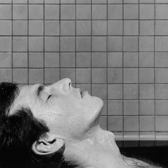 Close-up of a man in profile closing his eyes and leaning his head backward in front of a tile wall.