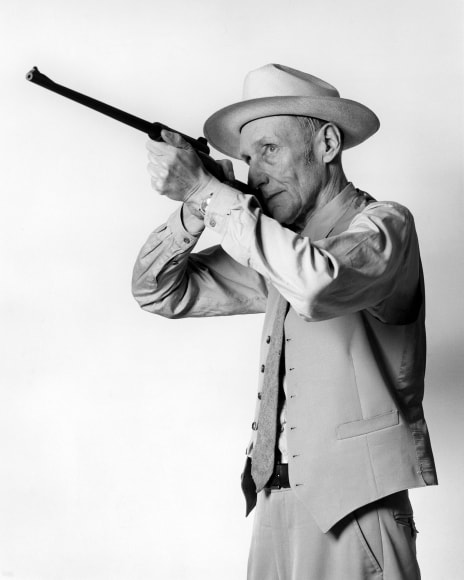 William Burroughs looking through a gun and wearing a suit.