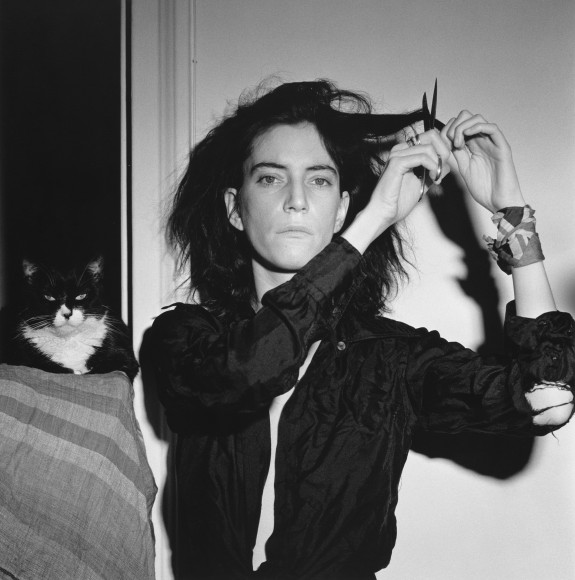Patti Smith facing camera, cutting her hair, with cat in background.
