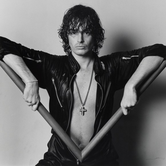 Man with damp, dark hair that hangs over his eyes wears a leather jumpsuit with chest exposed and holds a picture frame sample in front of him while gazing back at the viewer.