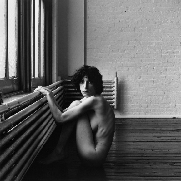 Nude Patti Smith with dark tousled hair sitting curled up in profile while holding on to a radiator by a window in a room with dark hardwood floors and white brick walls. Her head is turned to look directly at the viewer.