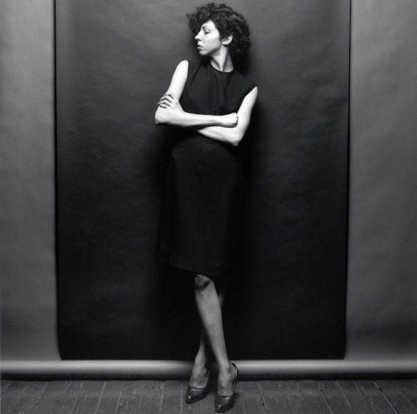 Woman with black sleeveless dress standing with arms crossed and head turned to the side in front of a geometric backdrop.