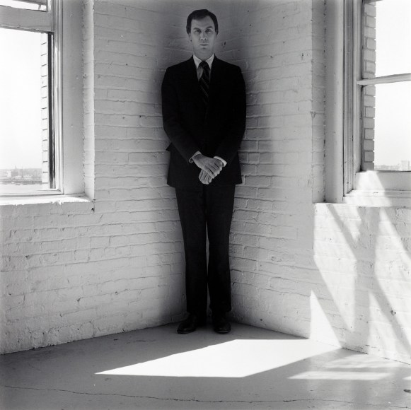 Robert Wilson wearing a suit standing in the corner of a white room.