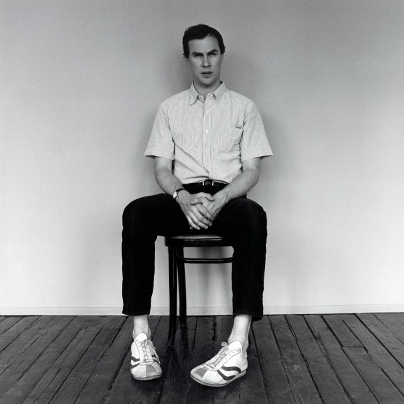Robert Wilson seated in chair with hands folded.