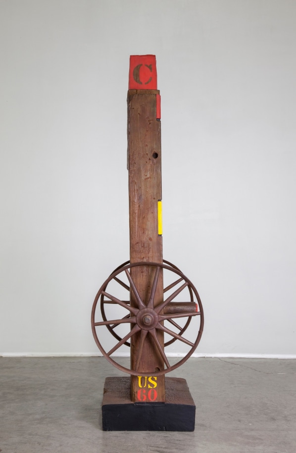 Left profile view of the bronze herm Hole. At the bottom of the left side &quot;US,&quot; appears in yellowed stenciled letters above &quot;60,&quot; stenciled in red. A wheel is attached to the side just above the text.