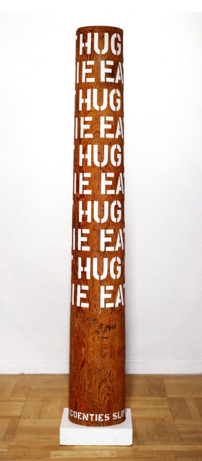 A column with ten rows of the words eat, hug, and die in gesso stenciled letters wrapping around the upper three quarters of the sculpture.