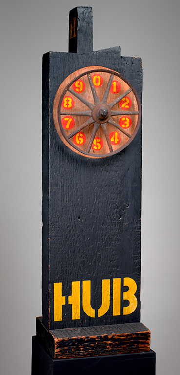 A 44 by 12 1/2 by 11 inch sculpture consisting of a wooden beam with a tenon on a wooden base. The sculpture is painted black, and its title, &quot;Hub,&quot; is painted in yellow stenciled letters across the bottom of the work. An iron and wood wheel has been affixed to the top front of the sculpture. The area behind the wheel has not been painted black, instead a yellow number from 0 to 9 in a red circle, has been painted between each spoke of the wheel, withe the numbers going clockwise.