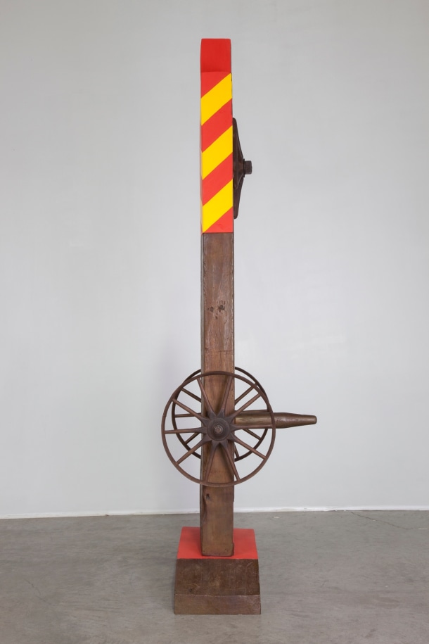 The left profile view of the herm Chief. Red and white danger stripes are visible in the top third of the sculpture, and a wheel is attached to the side, in the lower third of the work.