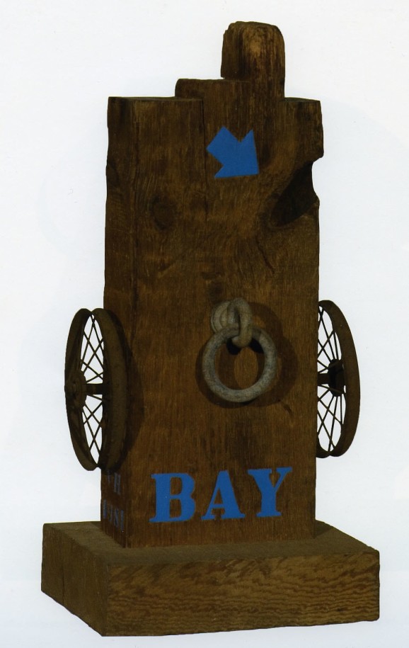 A 29 1/2 inch high sculpture consisting of a wooden beam with a haunched tenon. The work's title, Bay, is painted in blue letters across the lower front of the work, above the base. A small iron wheel is affixed on each side, and an iron ring on the front center. Above this is a painted blue arrow.