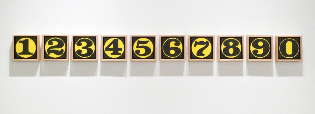 One Through Zero is a painting consisting of ten six inch square panels. Each panel consists of a different black numeral within a yellow circle against a black background. In this image the panels are arranged in a single horizontal row, starting with one and ending with zero.