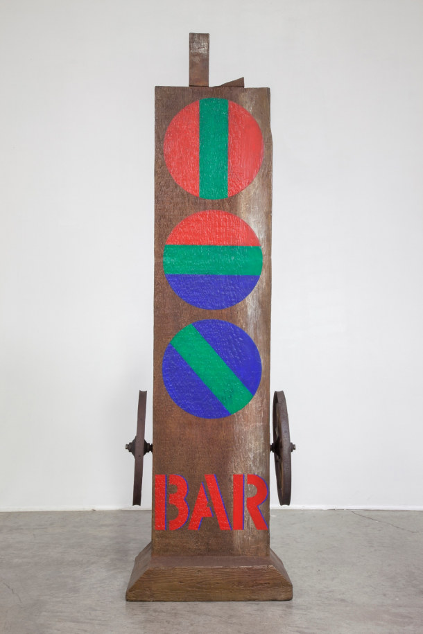A 58 5/8 by 15 by 12 5/8 inch painted bronze sculpture consisting of a beam with a haunched tenon, on a base. The sculpture's title, &quot;Bar,&quot; is painted in red stenciled letters across the bottom front of the work. Occupying the space from few inches above the title to the top of the sculpture are three painted red circles. The top one is red with a green vertical stripe down the center, the middle one consists of red, green and blue horizontal fields, and the bottom one is blue with a diagonal green stripe down the middle. A wheel has been affixed to the right and left sides of the sculpture, just above the title.