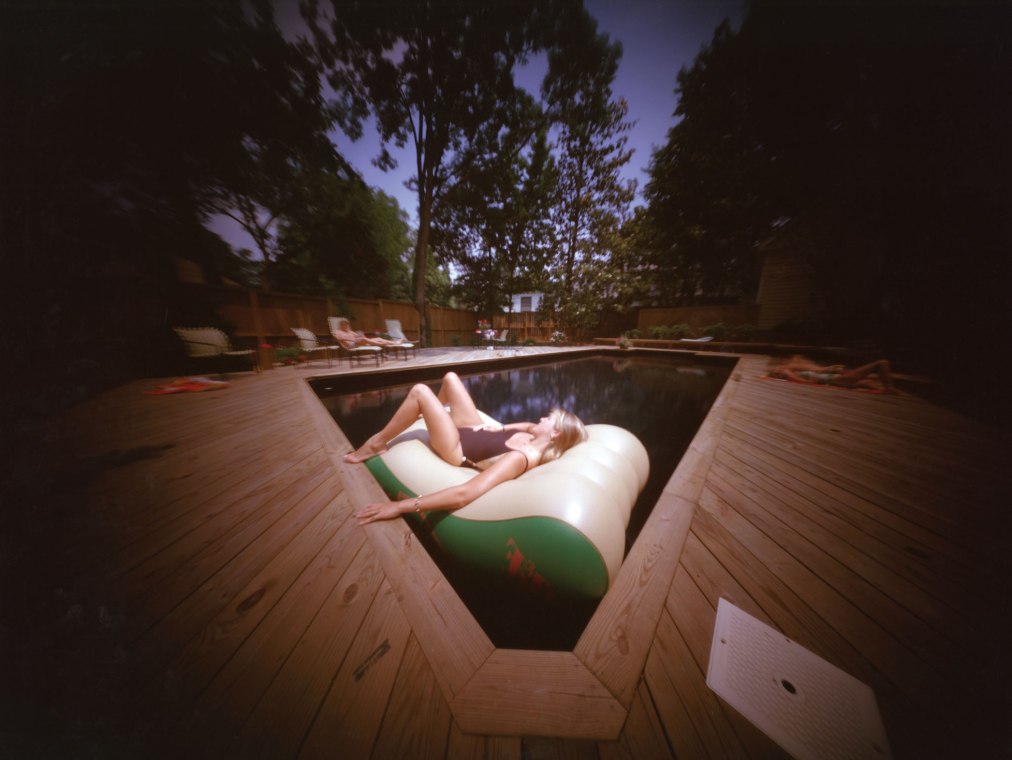 A woman sits leisurely on an inflatable in a pool surrounded by a wooden deck and nearby trees.