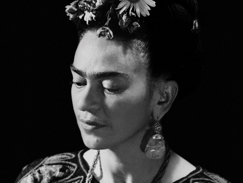 The artist Frida Kahlo is photographed in black and white as she wears traditional Mexican dress and flowers in her hair, while looking downwards.