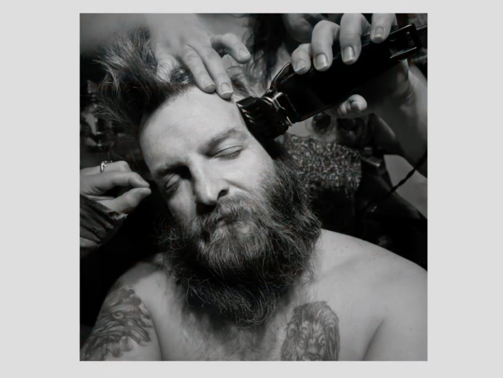 A bearded man has his long hair shaved in this black and white photo