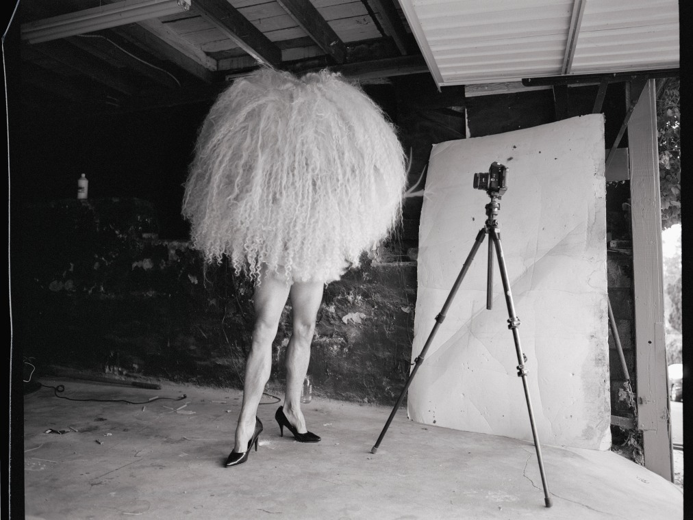 A man wearing high heels and a furry poof over his head and torso shows his naked legs to the camera.