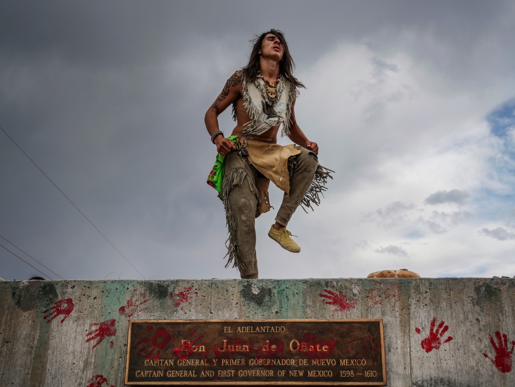 A young Native American man dances atop a memorial site and there are red hand prints all over the memorial as well.