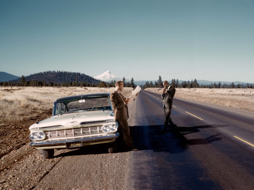 Two men, one white and one black, stand in a deserted roadway in nature alongside a 1950s car.