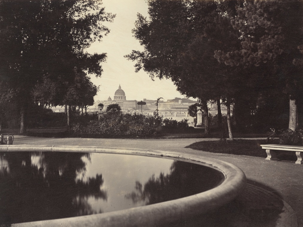 A view of Rome and a duomo in the distance from a park with a placid pool of water reflecting the trees.
