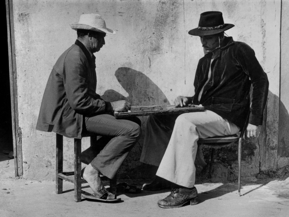 Two men sit at a table outside and play a game while wearing hats to protect themselves from the sun.