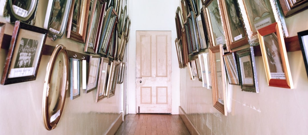Narrow hallway with wood floors and door at the end, hung with many framed pictures on both sides of the passage.