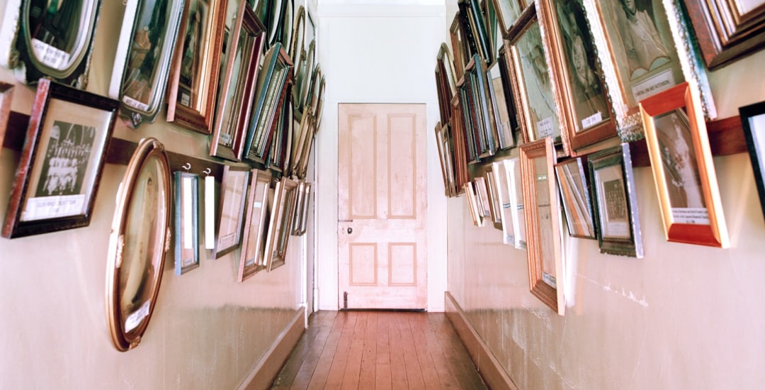 Narrow hallway with wood floors and door at the end, hung with many framed pictures on both sides of the passage.