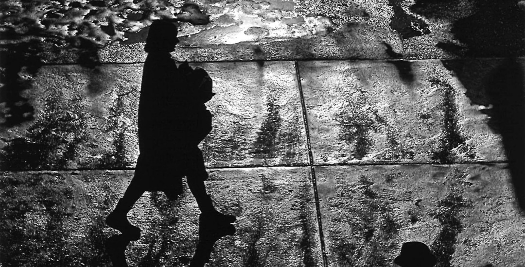 Black and white photo of a female pedestrian silhouetted abasing a shiny sidewalk that iw wet with melting snow and ice.
