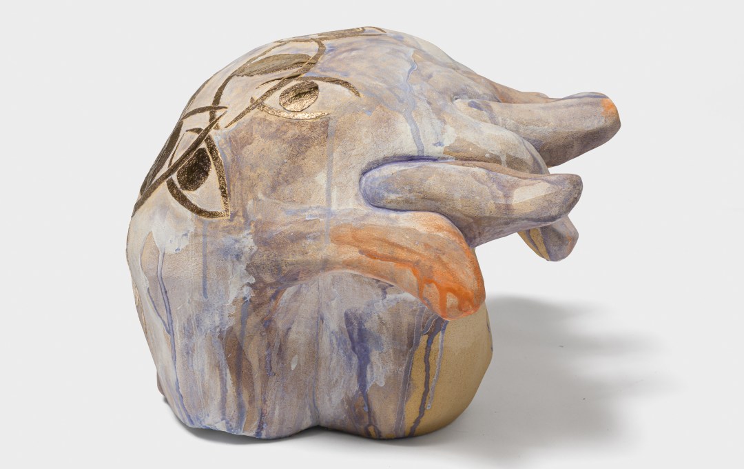 A ceramic sculpture of two hands interlocked with streaks of pale orange and blue glaze dripping down around golden eyes. 