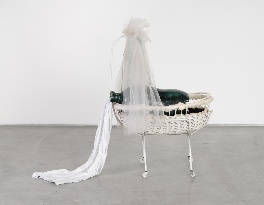 A sculpture by Daniel T. Gaitor-Lomack of a large glass vessel stuffed with a bedsheet in a baby's cradle.