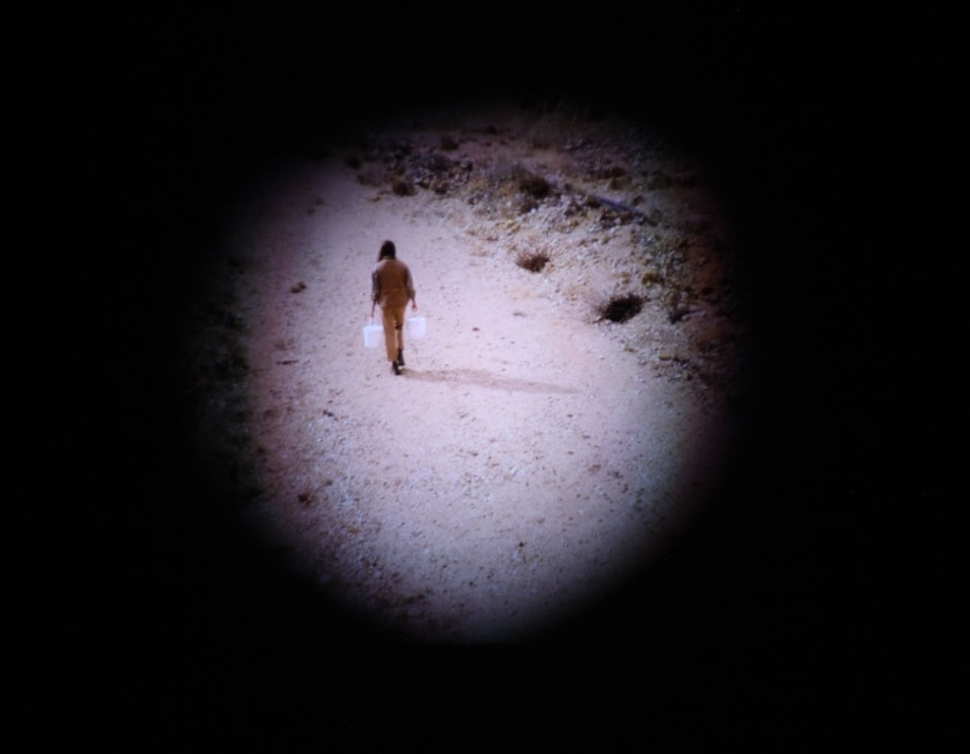 Video still showing the back of a woman as she walks up a rocky path in a quarry. She carries a bucket of clay in each hand, and they hang on either side of her as she walks. The video still is masked so that only a central circular portion of the image is visible; the rest of the frame is blacked out.
