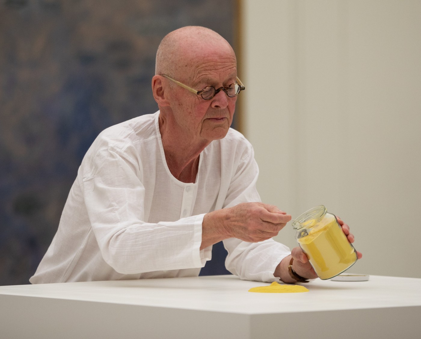 The artist, Wolfgang Laib, spoons yellow pollen from a jar onto a white pedestal, creating a mound of pollen.
