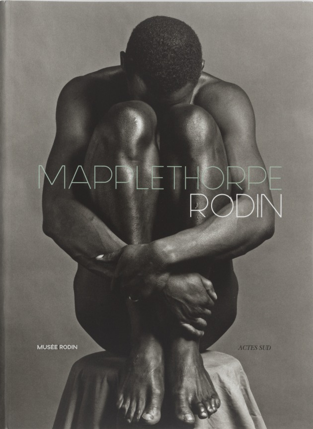 Ajitto sitting naked on a stool with his arms wrapped around his shins, and head against his knees.