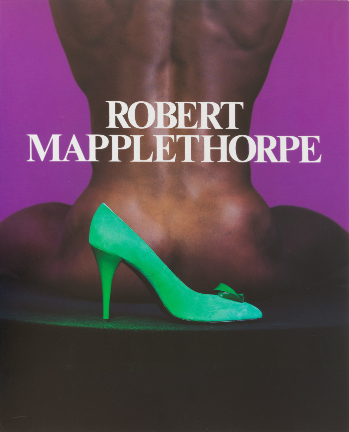 Color image of a bright green high-heeled shoe in front of a naked black man, seated and facing away from camera, against a purple background.