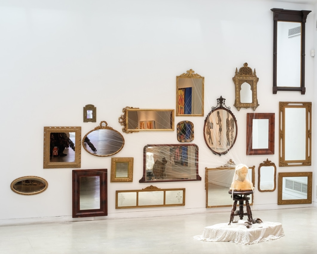 Rina Banerjee, She would be a vision of Beauty if only...her complexion was whitened! As if to not to see her and separately see part of her only not as human body as not yet whole, not yet made, as illness, as a single flaw with intent to take away, save it with this so to cure( vanish cream) her of ugliness, oldness, blemishes, freckles, moles and race deliver her as a sign of brightness, lightness, crowned beauty is a defining system. Where her otherness could marked, has been crowned, 2019,&nbsp;Antique mirrors, cotton thread, alabaster bust, antique lace, antique piano stool, mesh grocery bag, buffalo horns,&nbsp;Dimensions variable