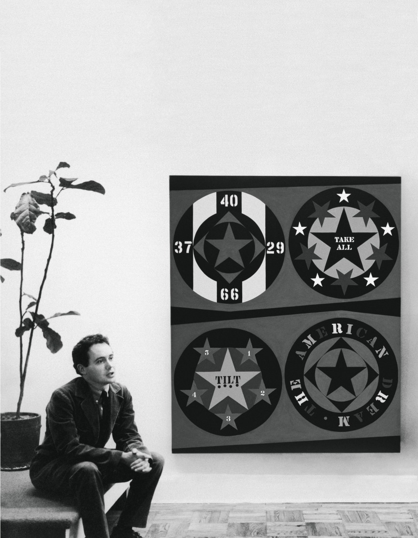 Black and white photograph of Robert Indiana, seated, next to his painting The American Dream, I
