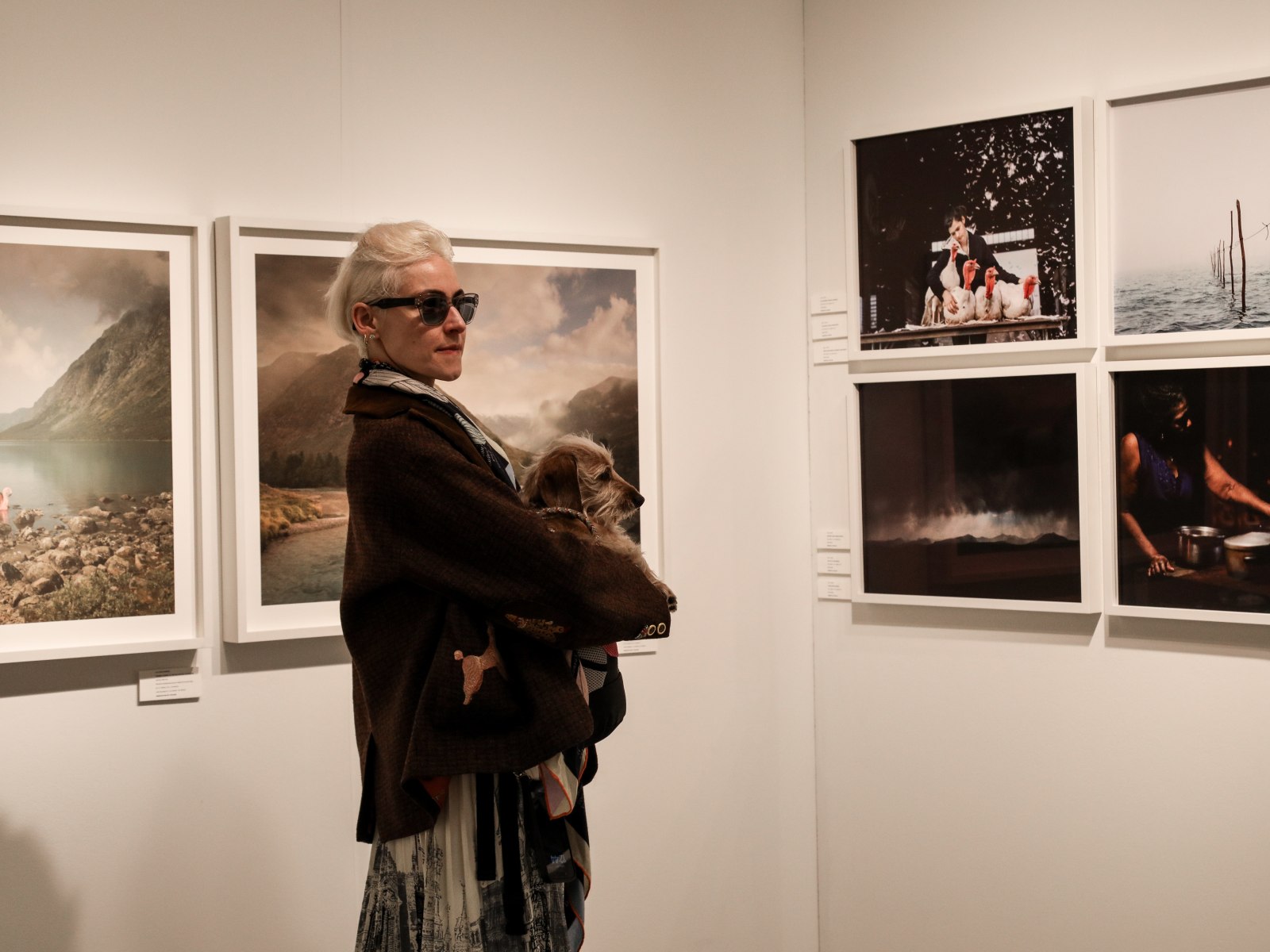 A blonde woman wears sunglasses and holds a dog while standing in a fair booth surrounded by color photographs.