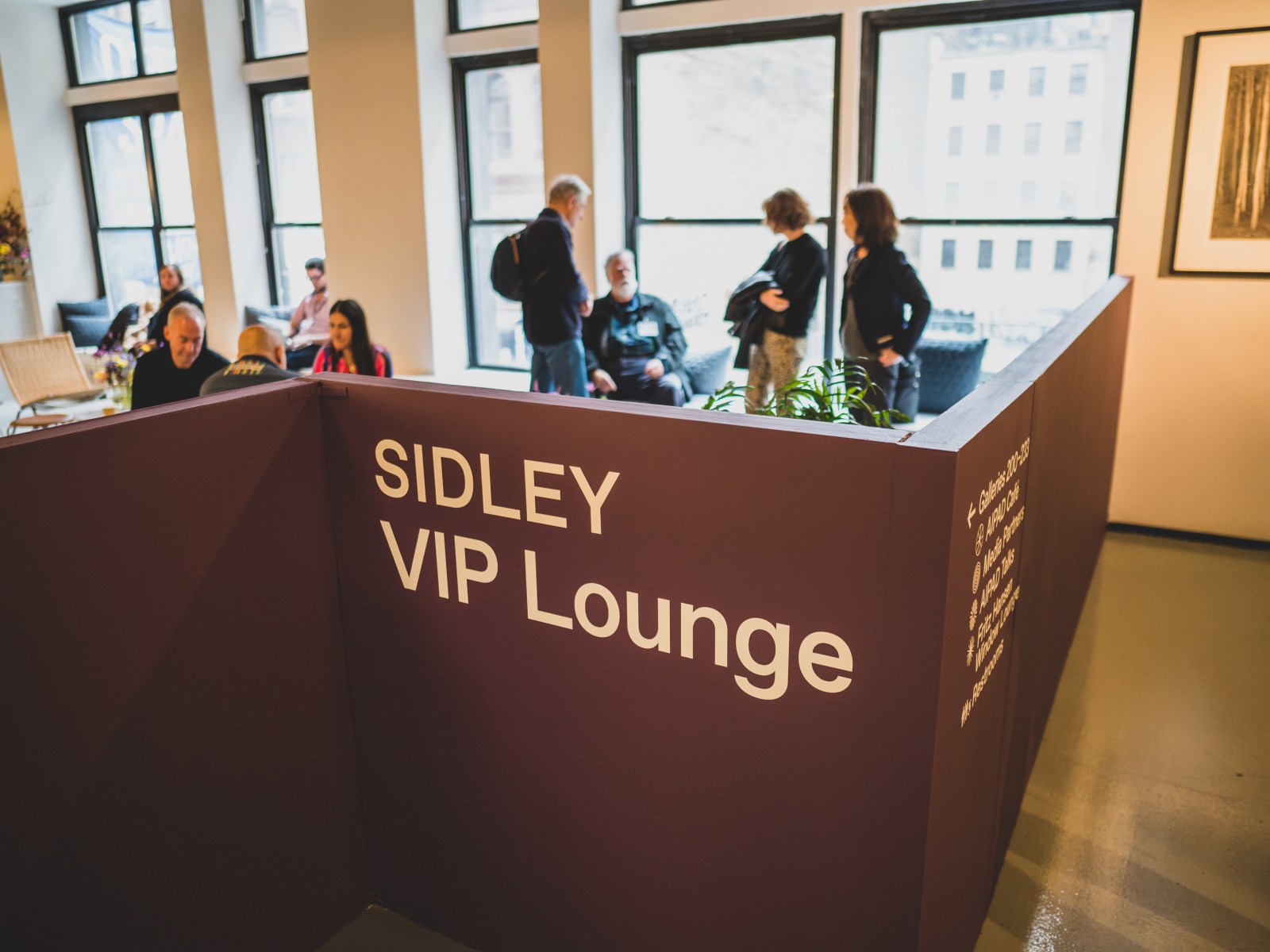 People sit and stand in the VIP Lounge at the 2023 Photography Show, with the Sidley name prominently featured.